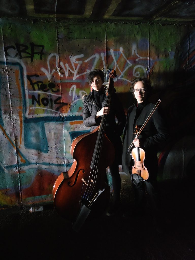 Press photo of Laura Dykes with her double bass and Jeffrey Young with his violin standing in a dark tunnel with lots of colorful graffiti on the wall including the phrase "Free Nois"