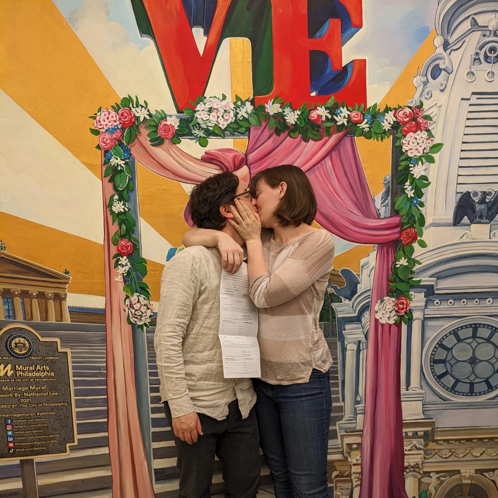 Jeff and Evie kissing in front of a mural depicting a wedding altar in front of famous Philadelphia landmarks while picking up their marriage license in Philadelphia City Hall!