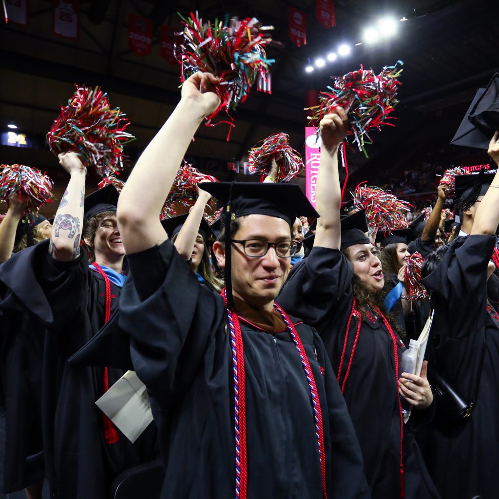 Jeffrey Young, wearing a graduation gown and honors cords, looking with a satisfied smile at the camera and holding aloft a pom-pom in a crowd with other graduating students at Rutgers